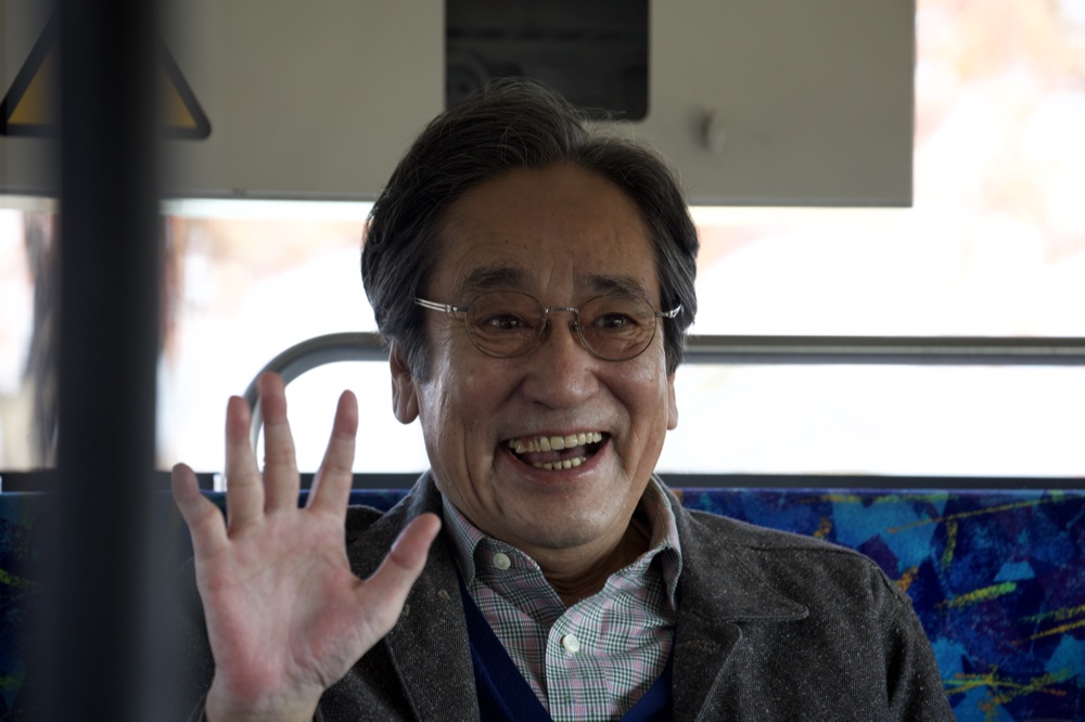 髙橋豊を演じるルー大柴。バスの車内で手を挙げて笑う。