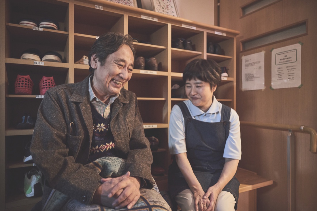 場面写真：施設の玄関で並んで腰かけ、笑い合う豊とスタッフ。